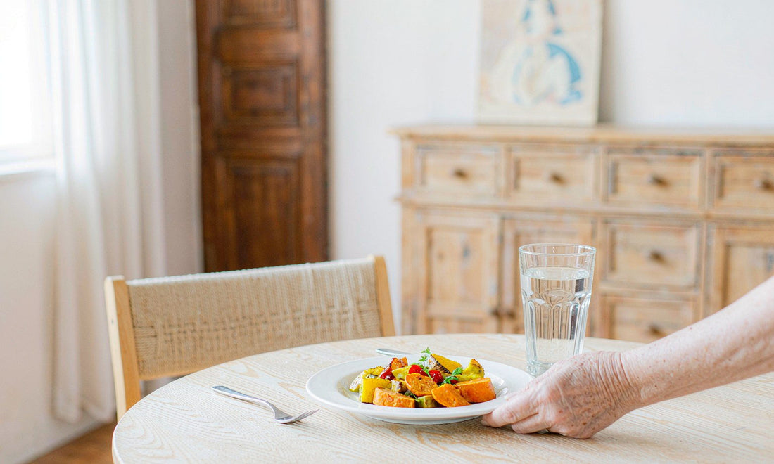 Fair-skinned hand of an older adult setting a plate of vegetables and water on a calm dining table