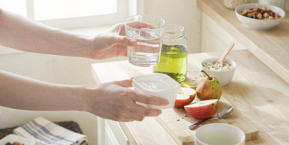 Fair-skinned hands holding water and a meal container on a bright kitchen counter for shift-worker wellness.