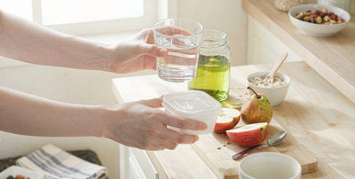 Fair-skinned hands holding water and a meal container on a bright kitchen counter for shift-worker wellness.