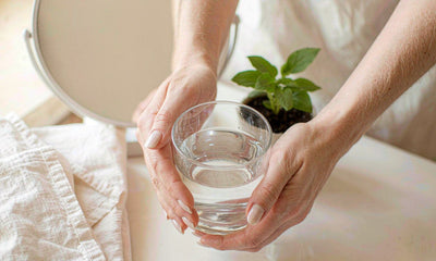 Hands lifting a glass of water from a white bathroom counter in morning light