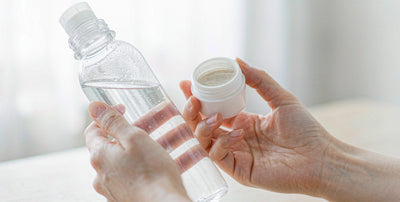 Fair-skinned hands holding a travel water bottle and a small jar on a bright table, no face visible