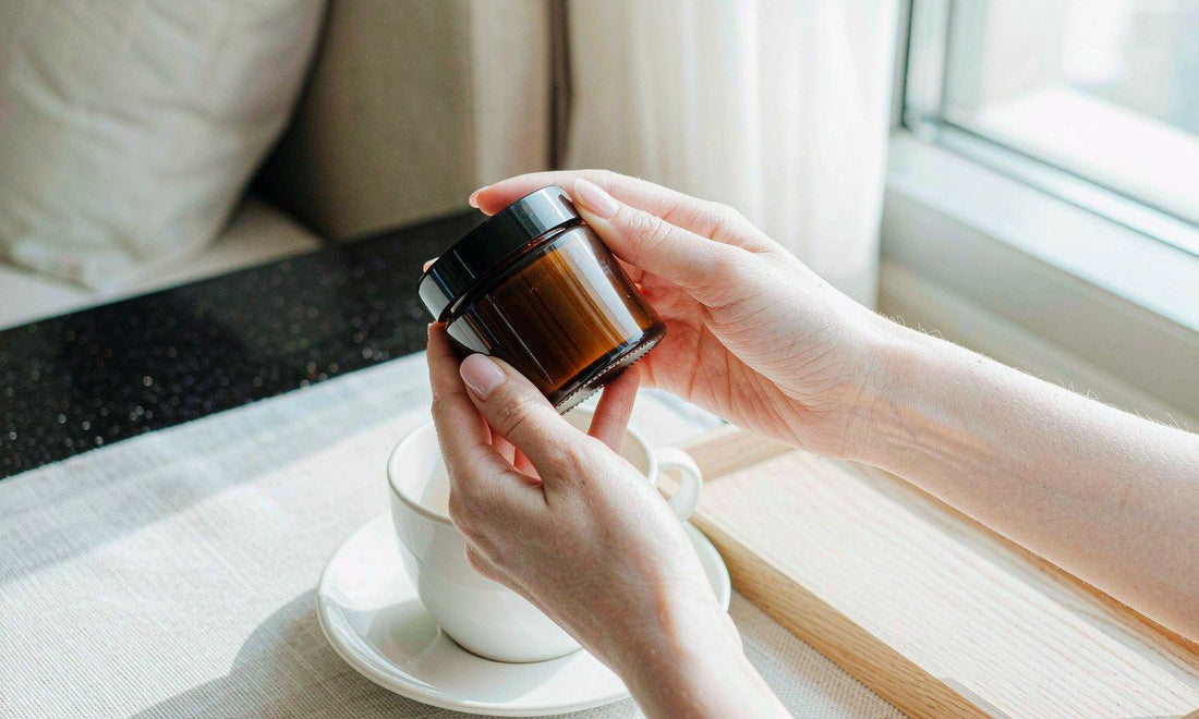 Fair-skinned hands holding a plain amber supplement jar beside a white coffee cup on a hotel table