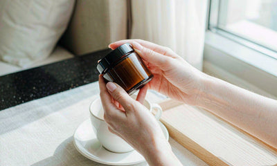 Fair-skinned hands holding a plain amber supplement jar beside a white coffee cup on a hotel table