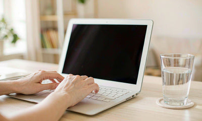Fair-skinned hands typing on a laptop beside a glass of water on a tidy home desk