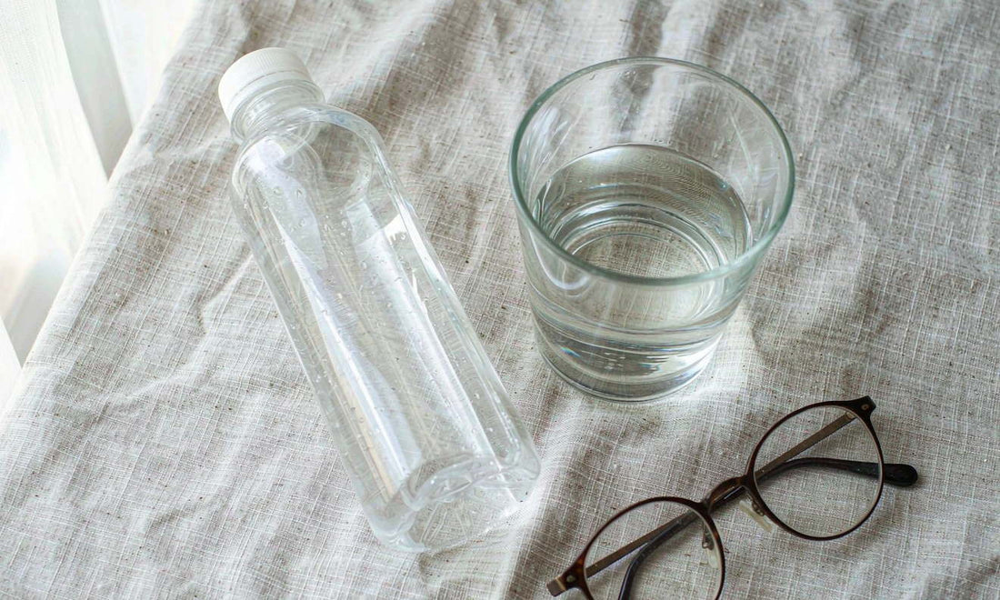 A plain unlabeled bottle beside a glass of water and reading glasses on a light linen cloth