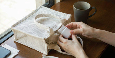 Fair-skinned hands placing a plain supplement jar into a tote at a sunny cafe