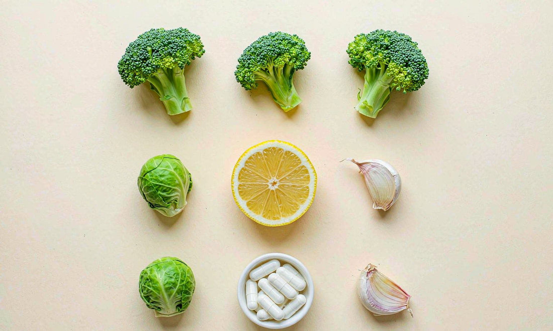 Flat lay of broccoli, Brussels sprouts, garlic, lemon and plain capsules on a pale cream surface