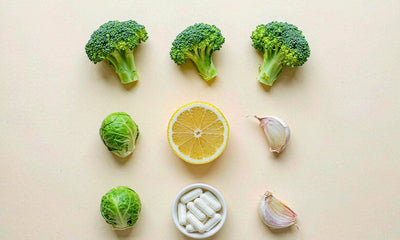 Flat lay of broccoli, Brussels sprouts, garlic, lemon and plain capsules on a pale cream surface